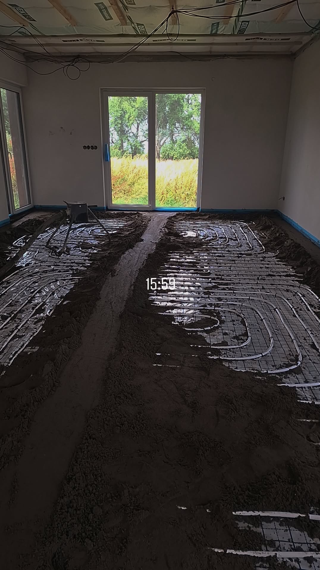 Instalacja ogrzewania podłogowego w surowym wnętrzu z widokiem na zieleń przez okno. Rury grzewcze ułożone na siatce, częściowo przykryte zaprawą. Widoczne przewody elektryczne.