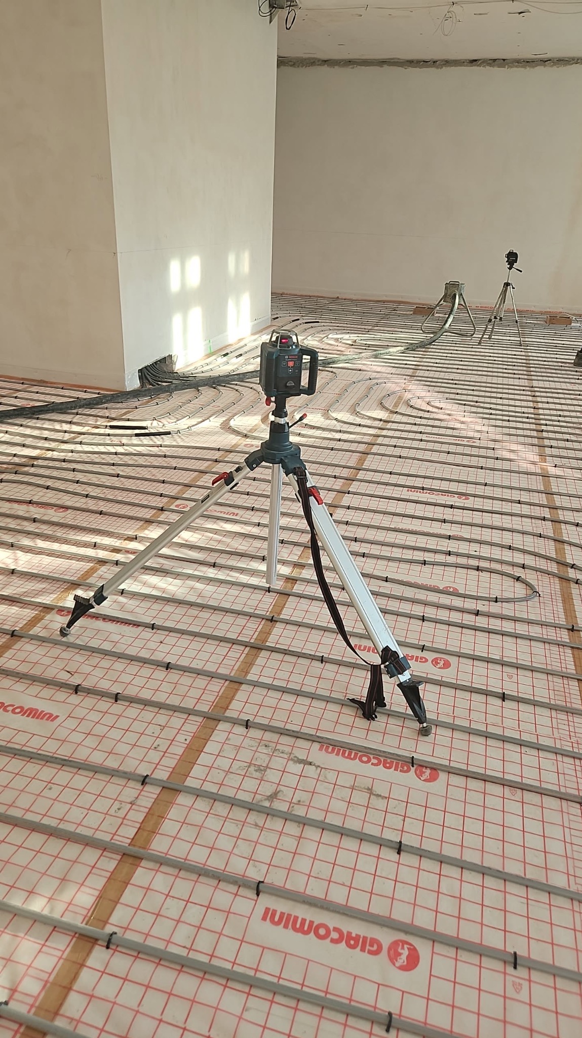 Rozłożone rury ogrzewania podłogowego na folii z siatką, poziomnica laserowa na statywie, widoczne elementy instalacji w surowym wnętrzu.