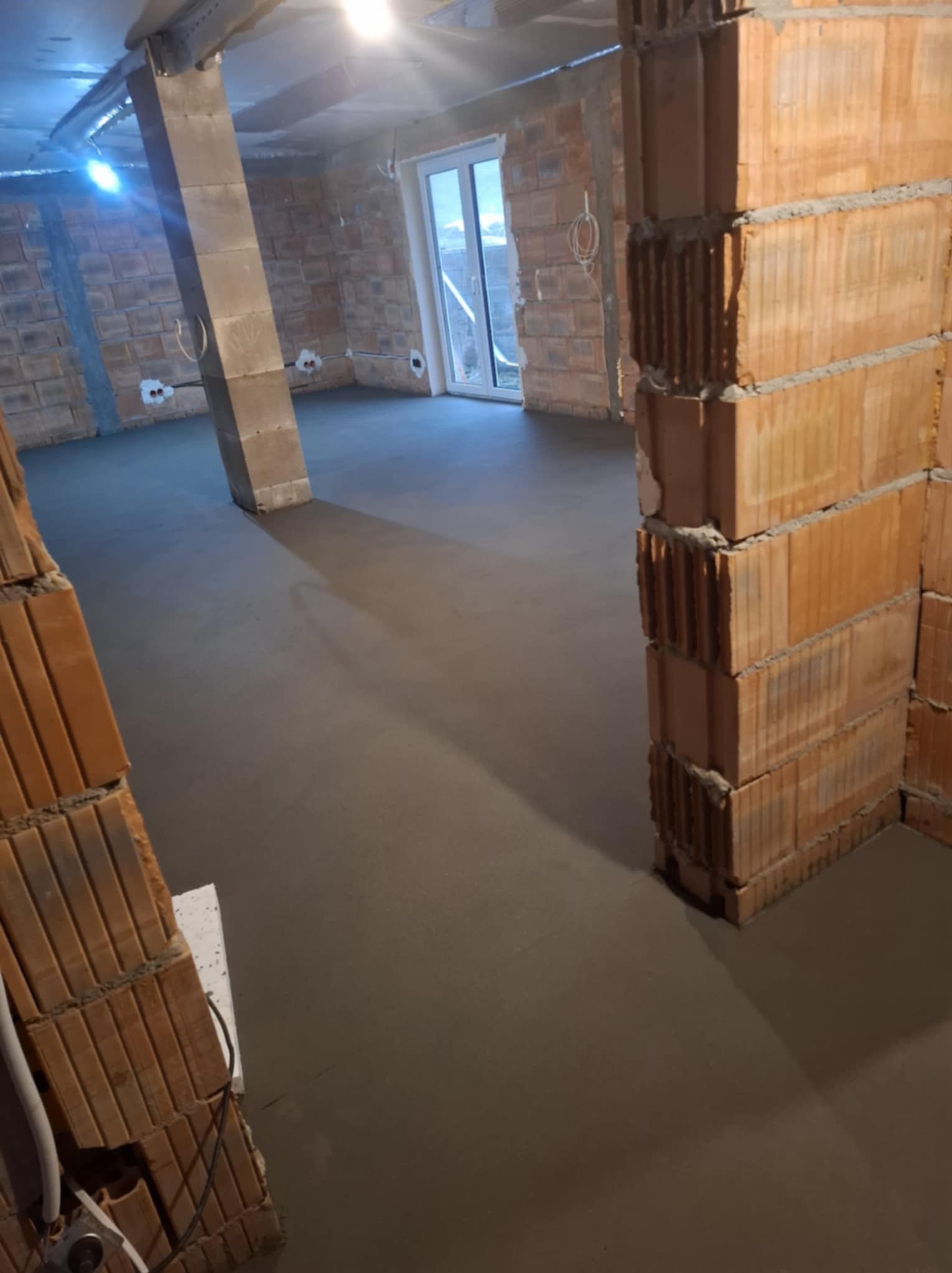 Świeża, gładka wylewka w surowym wnętrzu z ceglanymi ścianami i betonowym słupem. Widoczne instalacje elektryczne. Okno w tle.