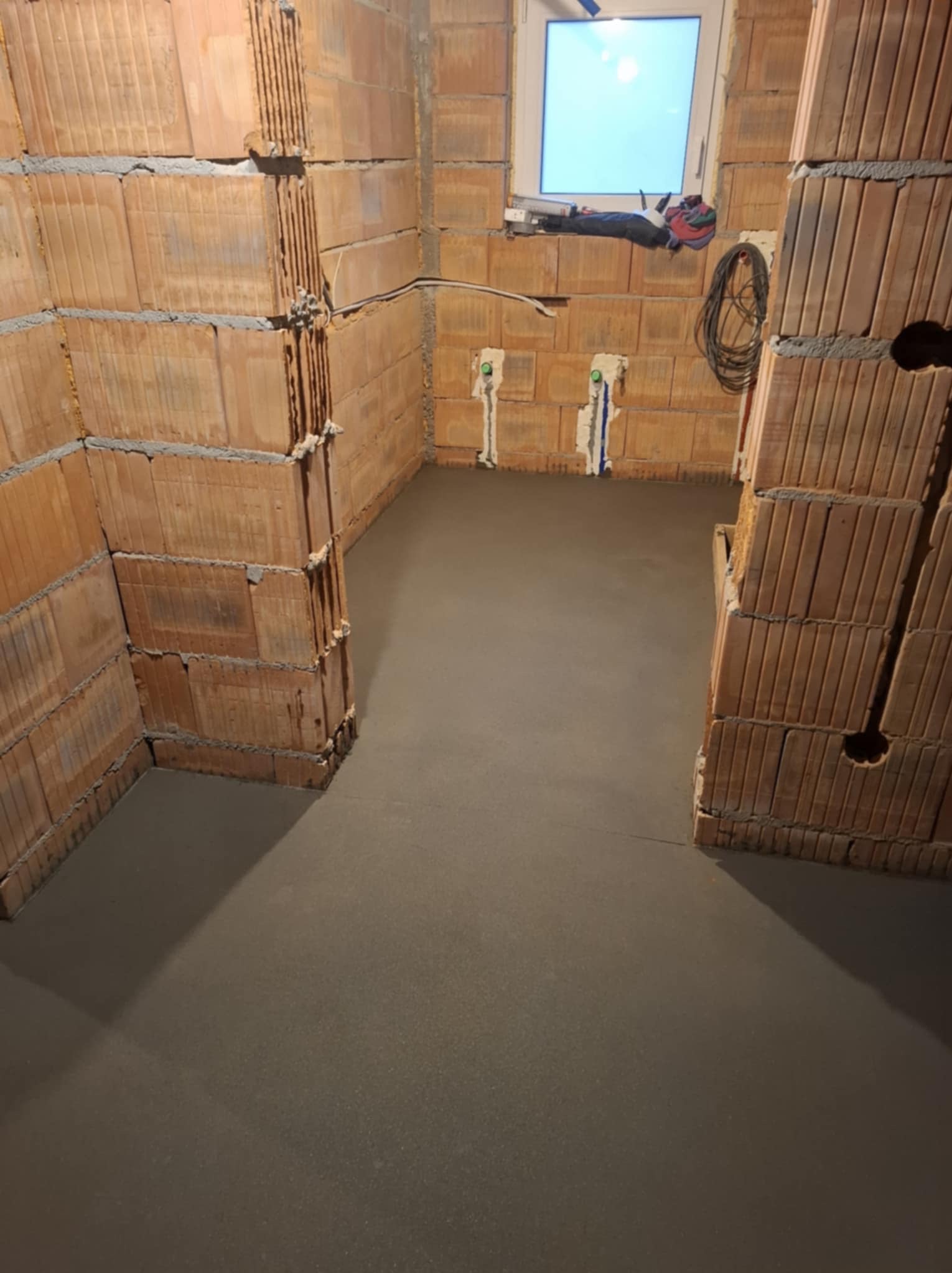 Gładka, szara wylewka w surowym wnętrzu z ceglanymi ścianami i widocznymi instalacjami. Okno w tle. Minimalistyczny, industrialny charakter.