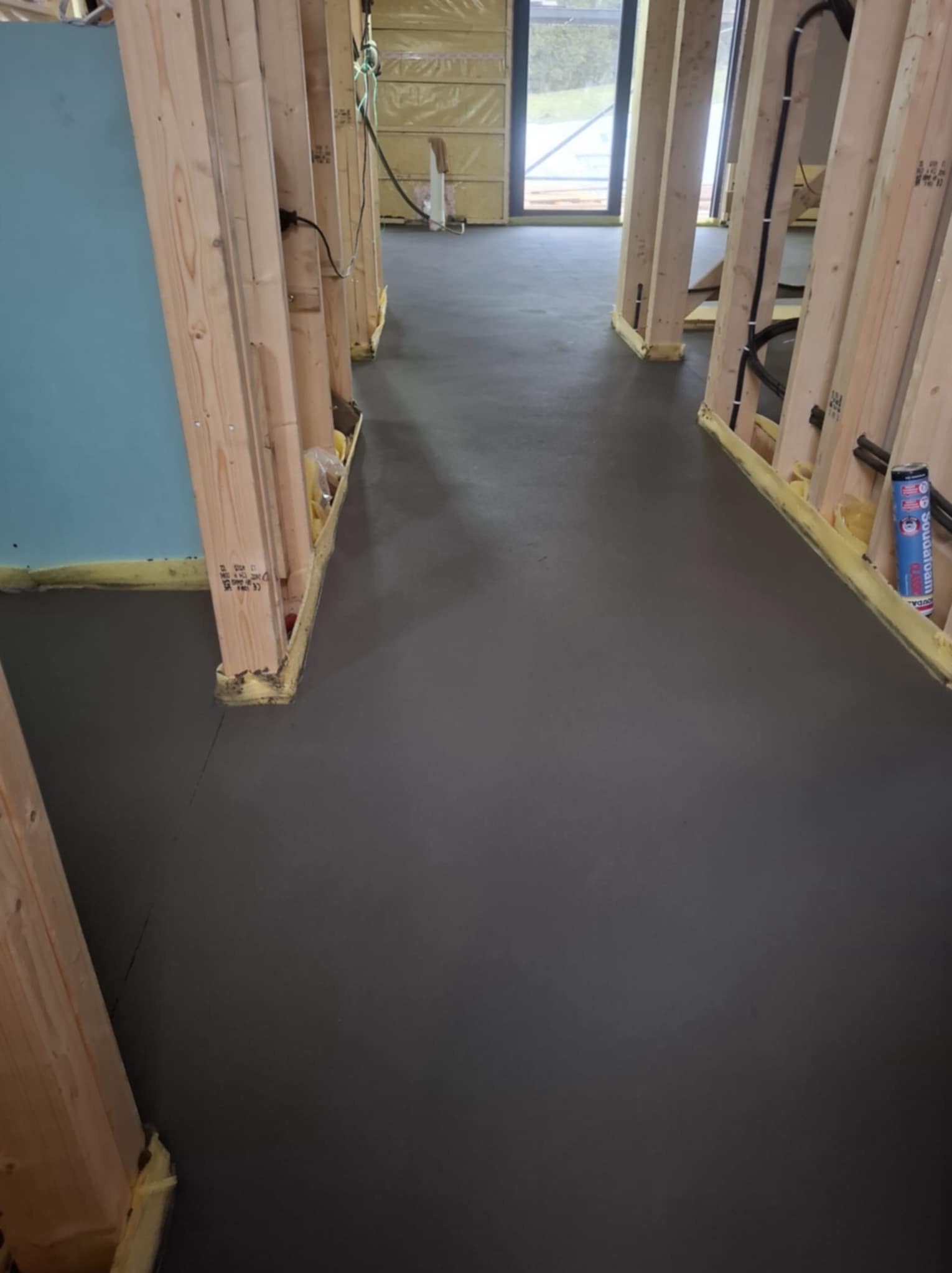 Świeża, szara wylewka wewnątrz budynku w trakcie budowy, widoczne drewniane elementy konstrukcyjne i izolacja. W tle okno i materiały budowlane.