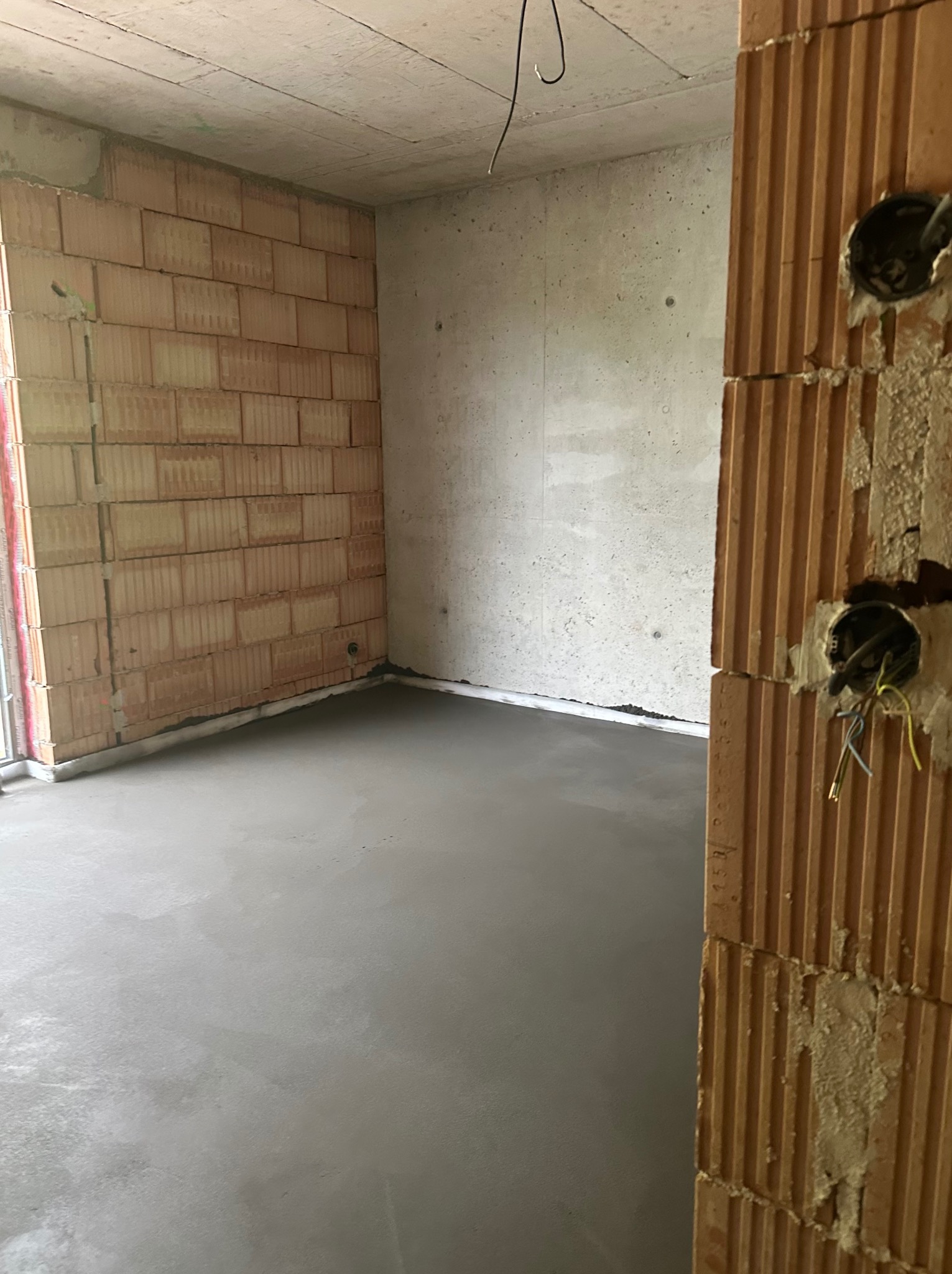 Świeża, gładka wylewka w surowym wnętrzu z ceglanymi ścianami i widocznymi puszkami elektrycznymi. Minimalistyczny beton w neutralnej tonacji.