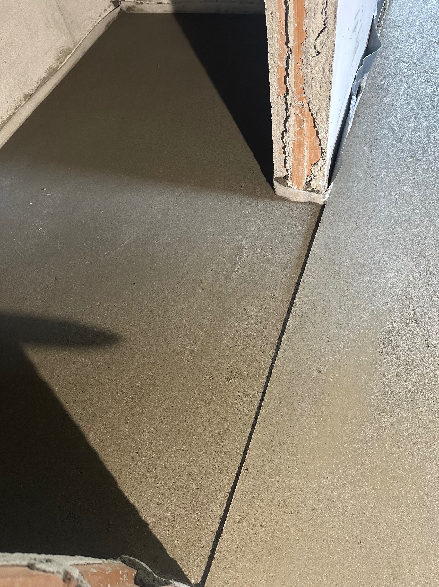 Świeżo wylana, gładka posadzka jastrychowa w pomieszczeniu, widoczna przy krawędzi ściany i progu drzwiowego z ceglaną obudową. Szara, jednolita powierzchnia.