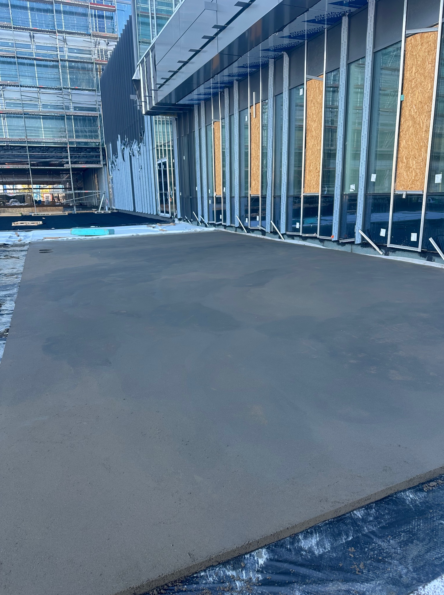 Świeżo wylana, gładka powierzchnia betonowa tarasu przy nowoczesnym budynku z oszkloną fasadą i rusztowaniami w tle. Widoczne detale wykończenia.