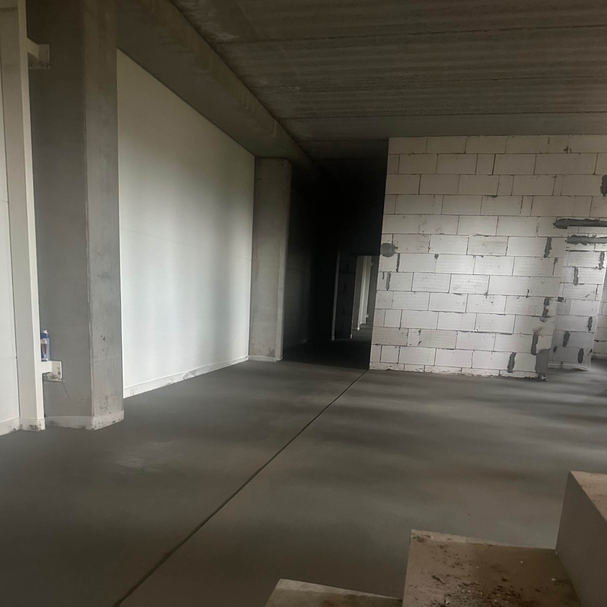 Wnętrze w trakcie budowy z gładką, szarą posadzką. Ściany z białych bloczków i surowego betonu. W tle widoczne ciemne przejście do kolejnych pomieszczeń.