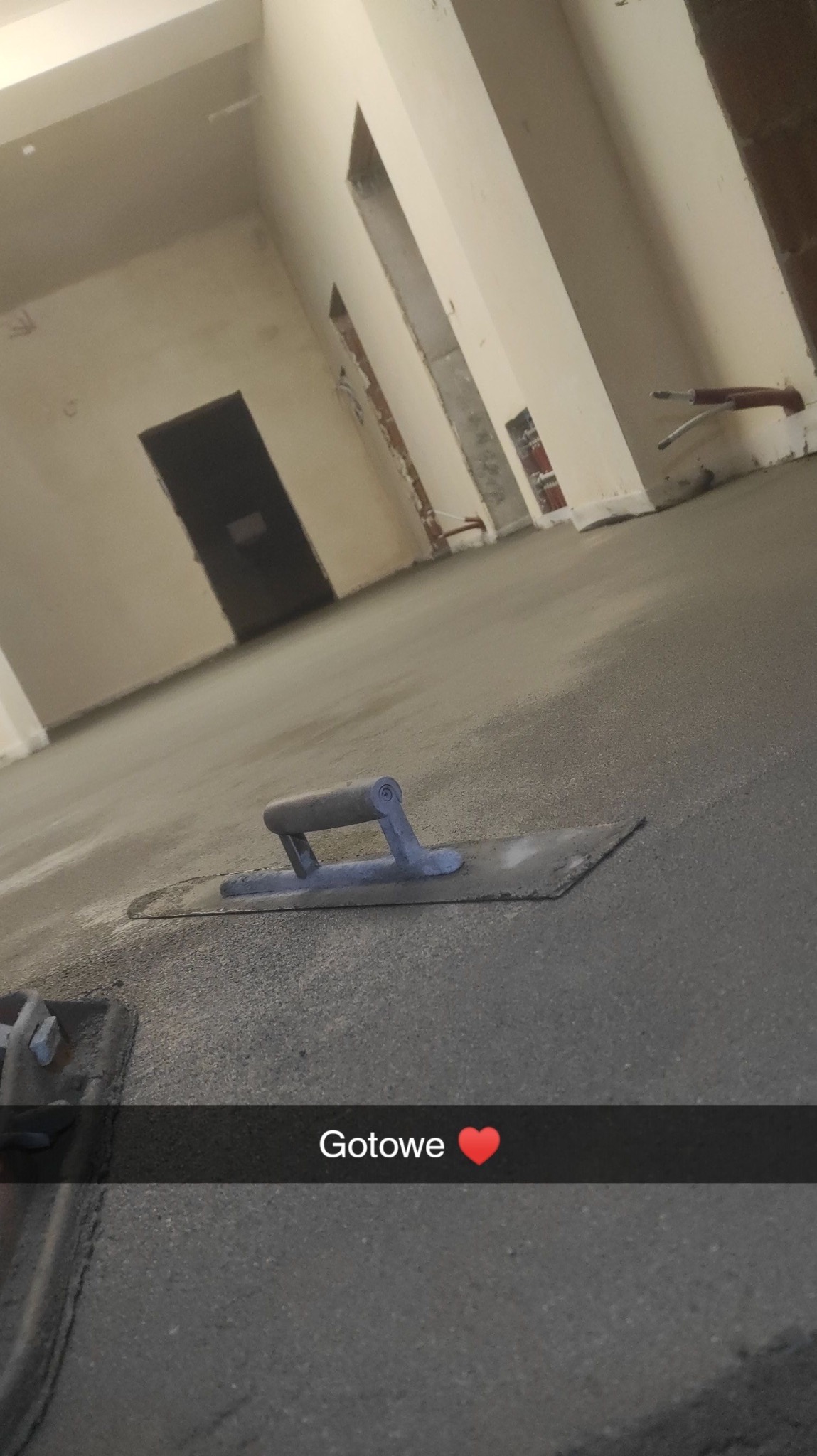 Wnętrze w trakcie prac wykończeniowych: świeżo wylana, szara posadzka z kielnią do wygładzania. W tle widoczne otwory drzwiowe i fragment instalacji.