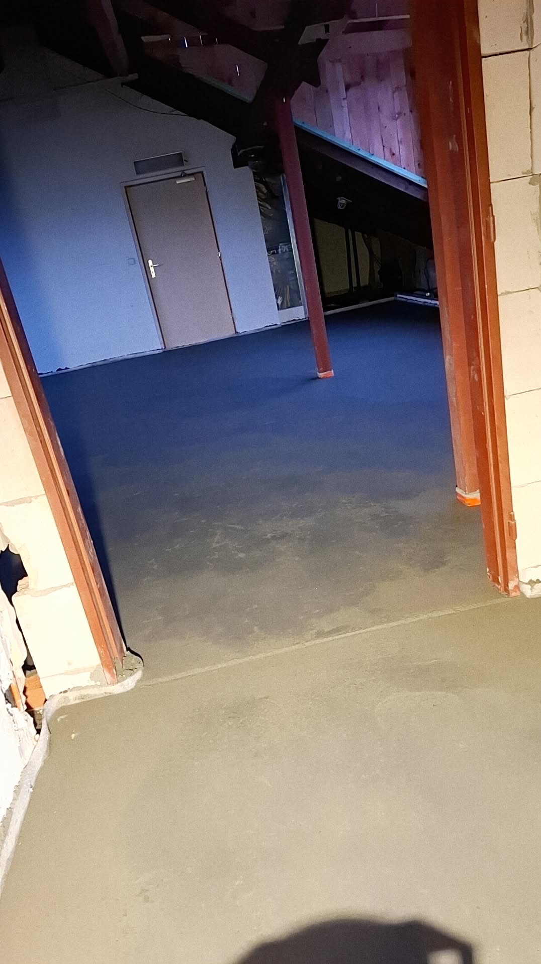 Świeżo wylana, gładka posadzka w surowym wnętrzu z widoczną konstrukcją dachu i fragmentem ściany z pustaków. Widoczne ślady po narzędziach i niedoskonałości w strukturze.