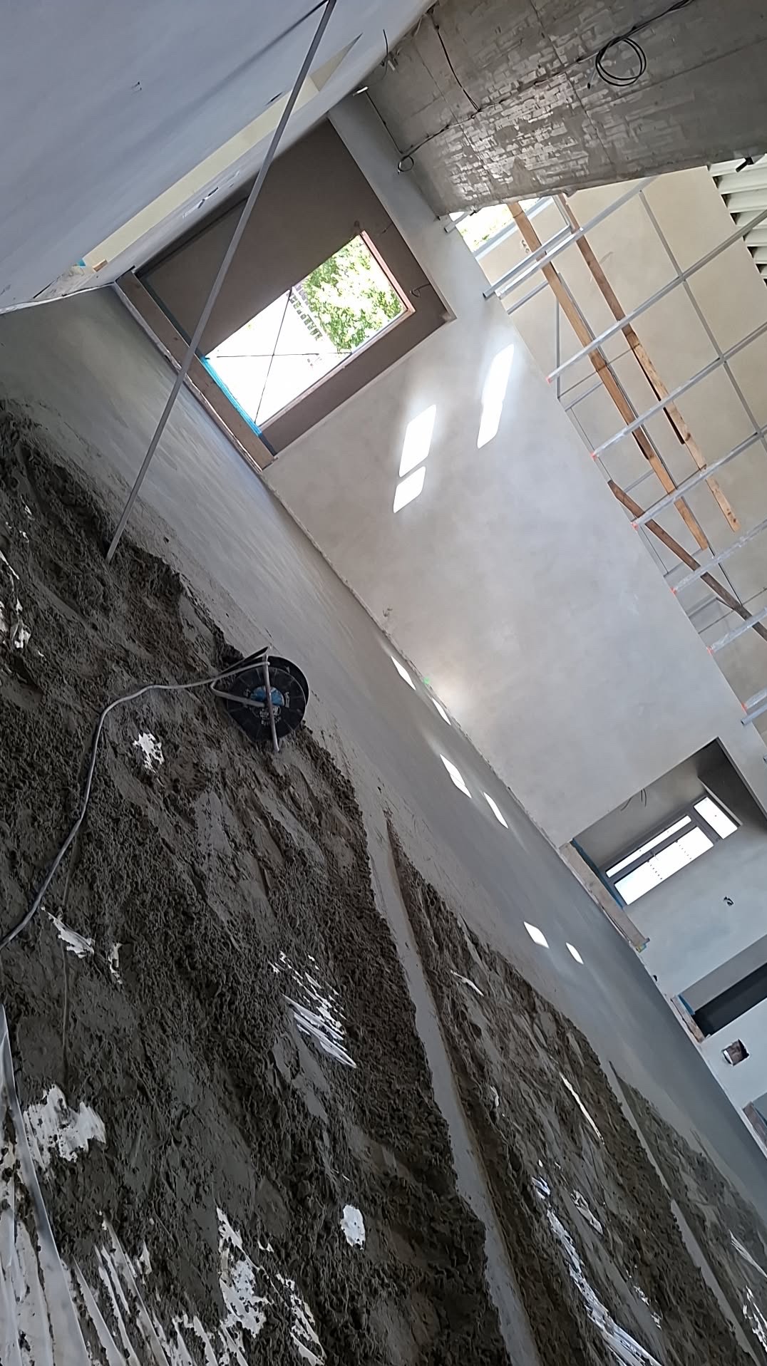 Świeżo nałożona, mokra wylewka na ścianie w surowym wnętrzu. Widoczne okno dachowe i elementy konstrukcji sufitu. Praca w toku, z użyciem specjalistycznego sprzętu.