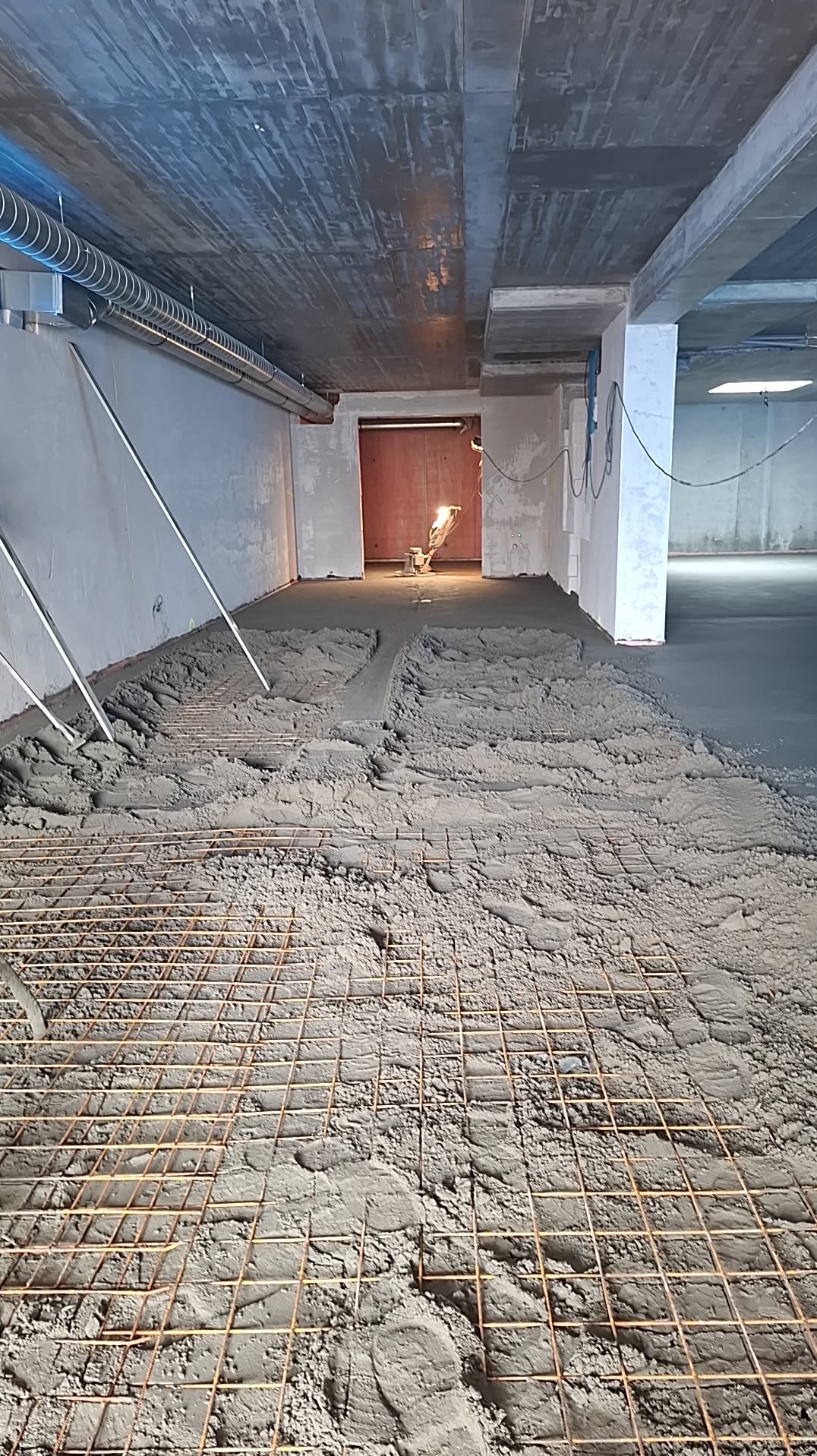 Wylewka jastrychowa zbrojona siatką stalową, świeżo po wylaniu. W tle widoczna maszyna do wylewania jastrychu i surowy betonowy strop. Szare kolory dominują.