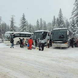 LenartTour.pl Przewóz Osób Paweł Lenart - Autokary na parkingu w zimowej scenerii, osoby z nartami i snowboardami. Śnieg pada, drzewa pokryte śniegiem. Przygotowania do wyjazdu na stok narciarski.