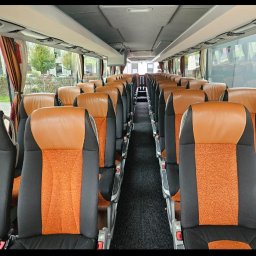 LenartTour.pl Przewóz Osób Paweł Lenart - Wnętrze autobusu turystycznego z rzędami foteli w kolorach brązowym, pomarańczowym i czarnym. Widok na przejście między siedzeniami, zasłony w oknach.