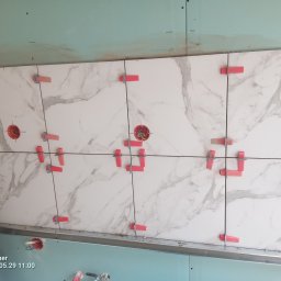 Usługi budowlane - Białe płytki imitujące marmur, ułożone na ścianie z użyciem czerwonych klipsów poziomujących. Widoczne puszki elektryczne w ścianie.