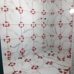 Usługi budowlane - Białe płytki imitujące marmur, ułożone w łazience, z czerwonymi klipsami poziomującymi. Widoczne otwory na instalacje. Oświetlenie halogenowe.