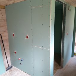 Usługi budowlane - Ściana z płyt gipsowo-kartonowych z widocznymi otworami na instalacje, drewniany sufit i podłoga, metalowa futryna drzwiowa. Etap prac wykończeniowych.