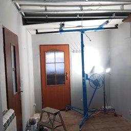 Usługi budowlane - Montaż sufitu podwieszanego: metalowa konstrukcja, podnośnik do płyt, drabina, drzwi. Widoczne narzędzia i materiały. Oświetlenie robocze.