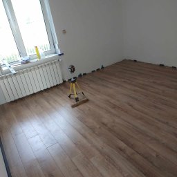 Usługi budowlane - Układanie paneli podłogowych w pokoju z użyciem poziomicy laserowej. Częściowo ułożona podłoga z brązowych paneli. Widoczne listwy przypodłogowe i okno.