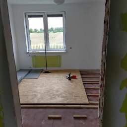 Usługi budowlane - Montaż podłogi: płyty OSB na legarach w pokoju z oknem na pole. Widoczne narzędzia i szczelina w ścianie. Etap prac przed finalnym wykończeniem.