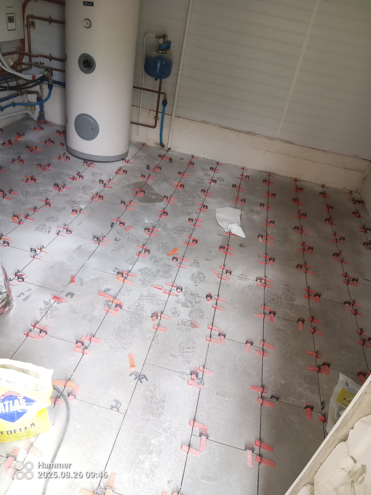 Szare płytki podłogowe w trakcie układania, z czerwonymi klipsami poziomującymi, widoczne ślady stóp i worek zaprawy Atlas Geoflex. W tle bojler i biała boazeria.