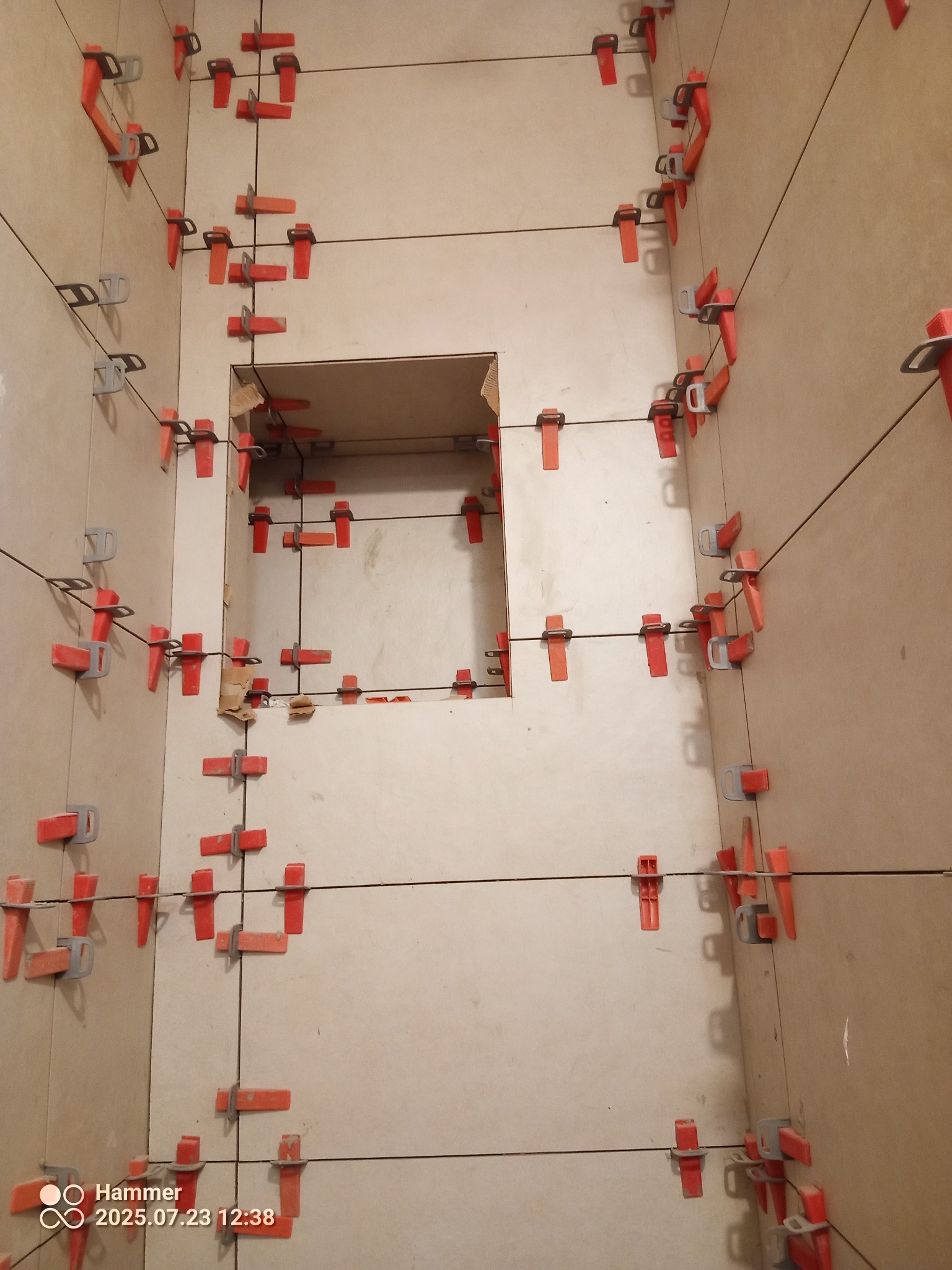 Ściana z jasnych płytek, w trakcie montażu, z użyciem czerwonych klipsów poziomujących. Widoczny otwór w ścianie. Ujęcie z bliska, z perspektywy podłogi.