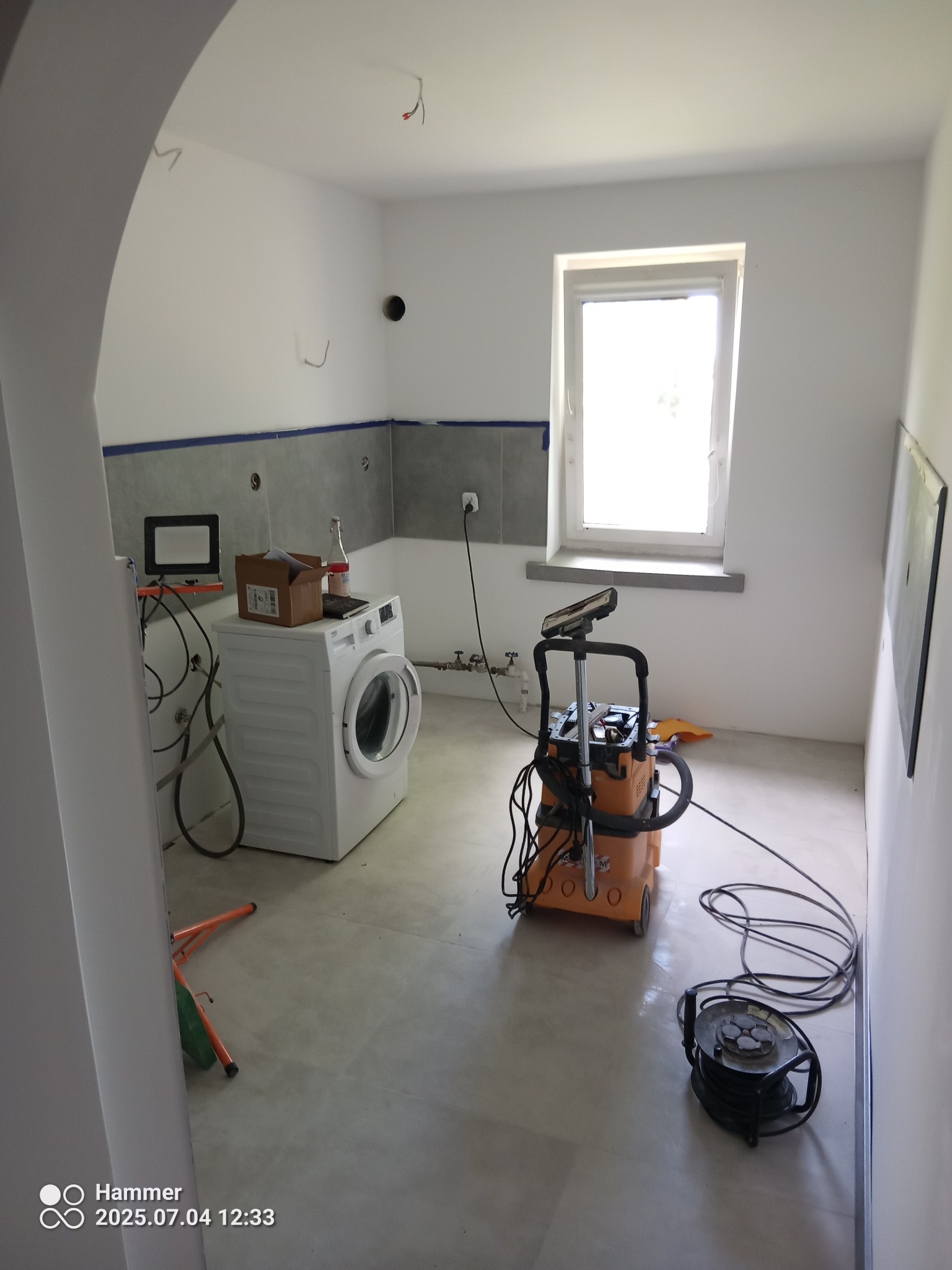 Wnętrze łazienki w trakcie remontu: częściowo położone płytki, pralka, odkurzacz przemysłowy i inne narzędzia. Widoczne niedokończone instalacje.