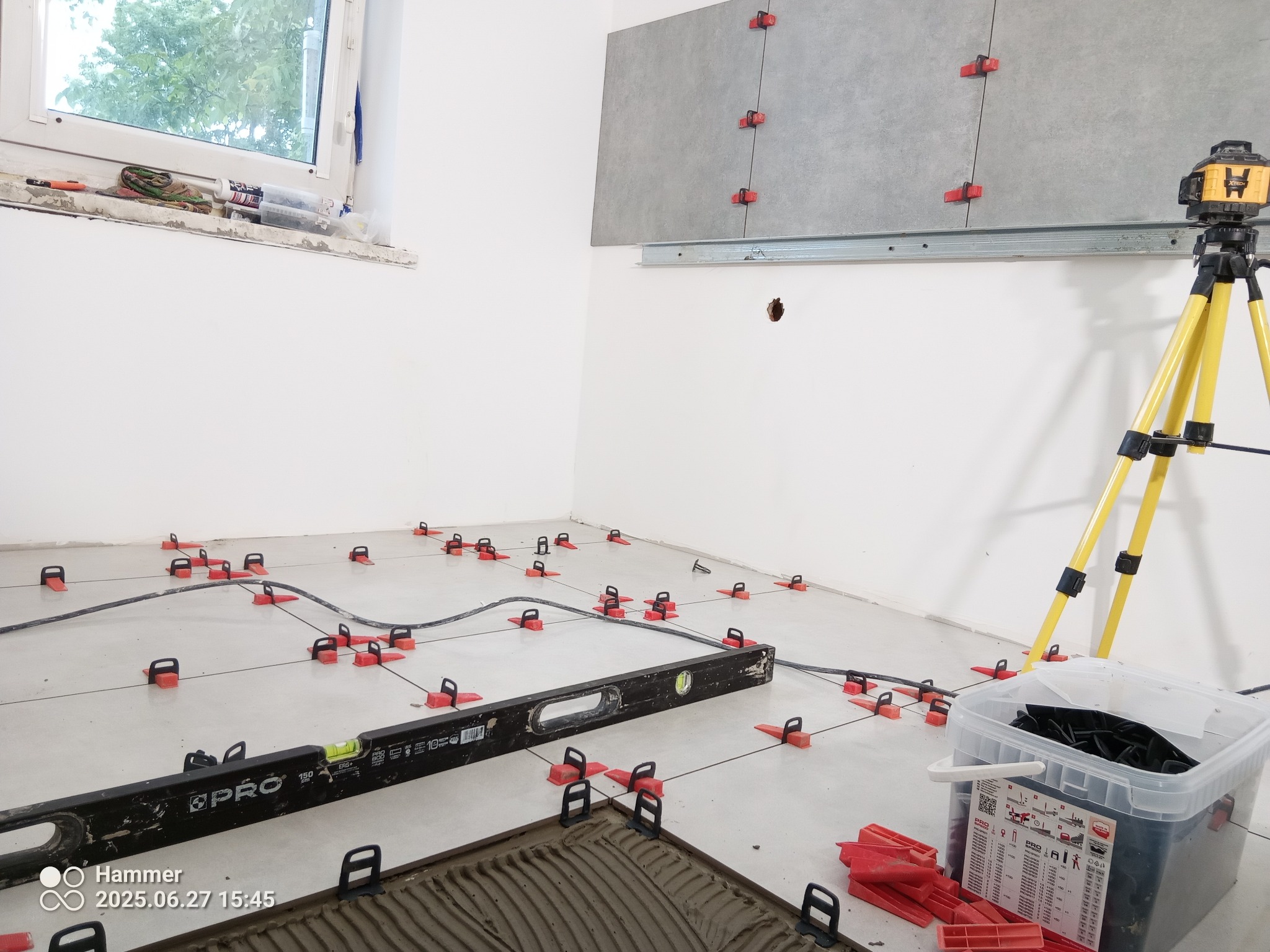 Proces układania płytek: poziomica, laser krzyżowy na statywie, system poziomowania płytek na podłodze i ścianie. Widoczne narzędzia i materiały.