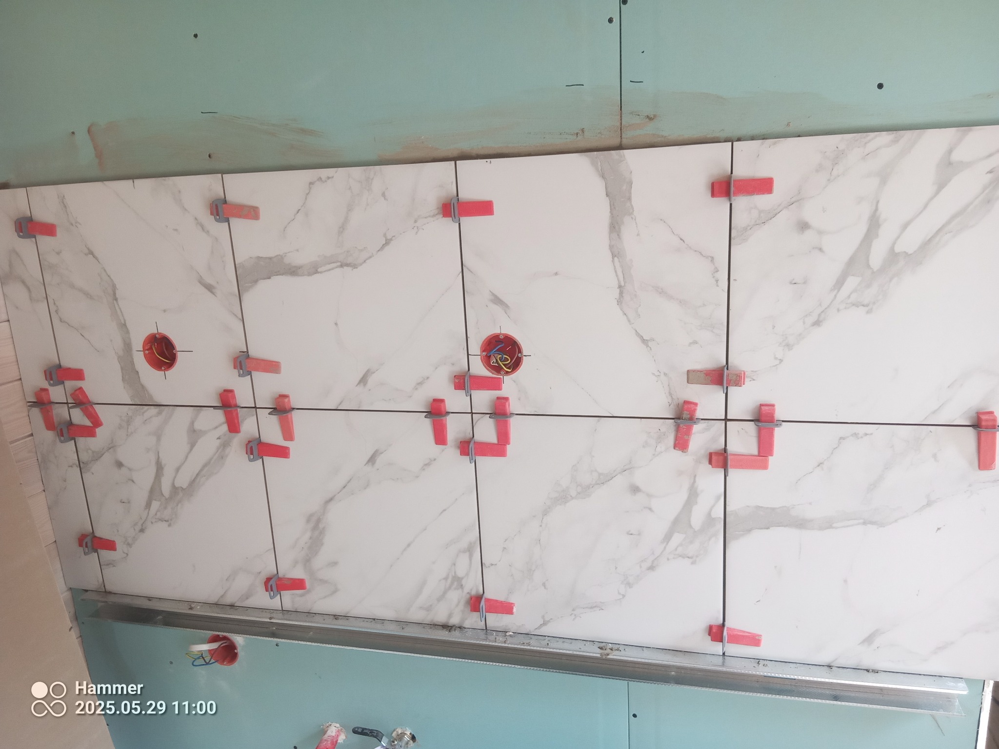 Białe płytki imitujące marmur, ułożone na ścianie z użyciem czerwonych klipsów poziomujących. Widoczne puszki elektryczne w ścianie.