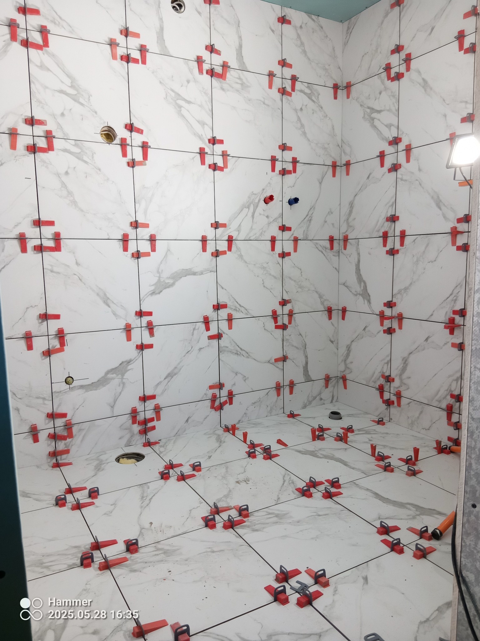 Białe płytki imitujące marmur, ułożone w łazience, z czerwonymi klipsami poziomującymi. Widoczne otwory na instalacje. Oświetlenie halogenowe.