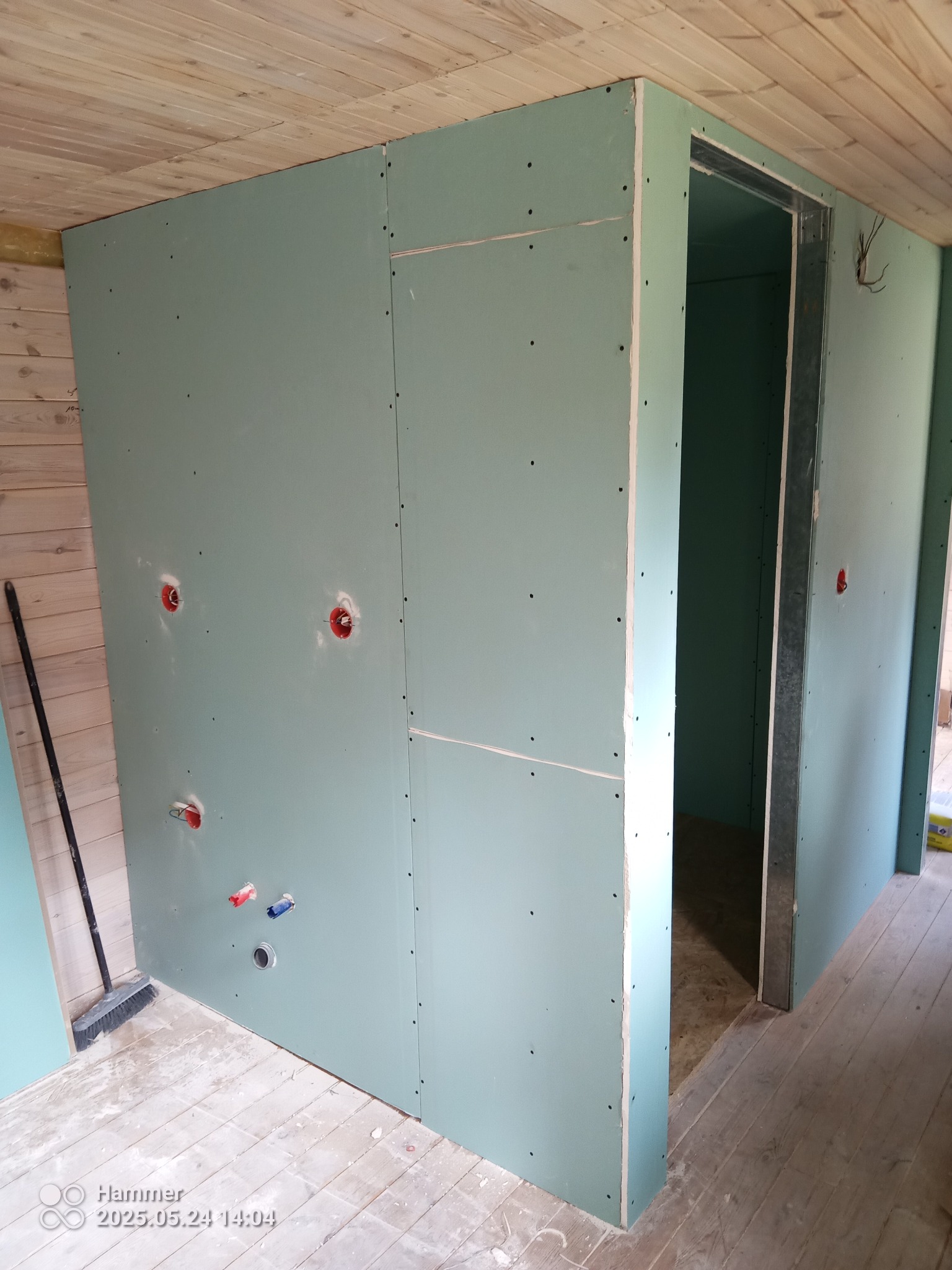 Ściana z płyt gipsowo-kartonowych z widocznymi otworami na instalacje, drewniany sufit i podłoga, metalowa futryna drzwiowa. Etap prac wykończeniowych.