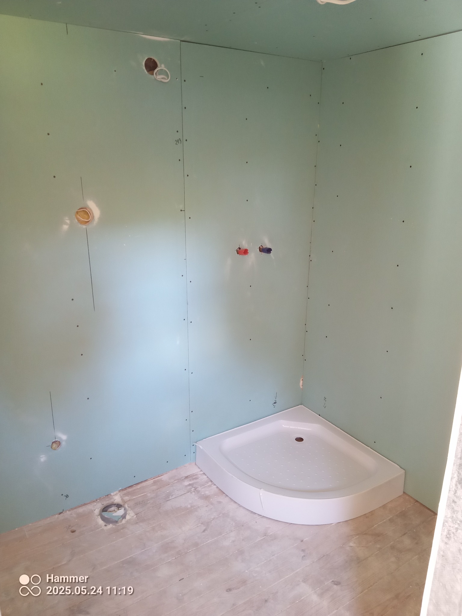 Narożny brodzik w surowej łazience z płytami GK, widoczne instalacje wodne i kanalizacyjne. Etap przygotowawczy przed wykończeniem.