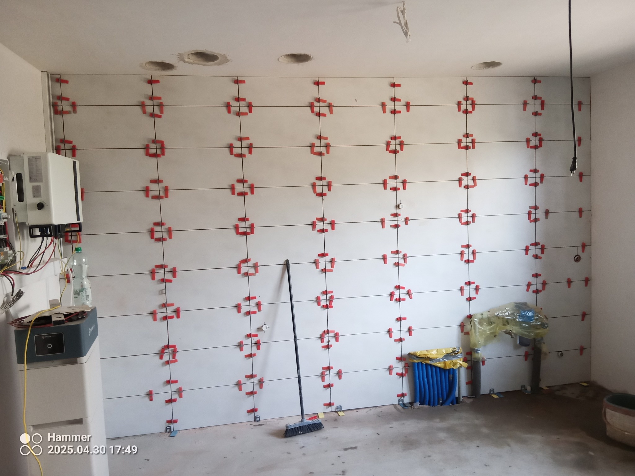 Ściana z białych płytek, w trakcie układania, z czerwonymi krzyżykami dystansowymi. Widoczne instalacje i narzędzia. Etap prac wykończeniowych w pomieszczeniu.