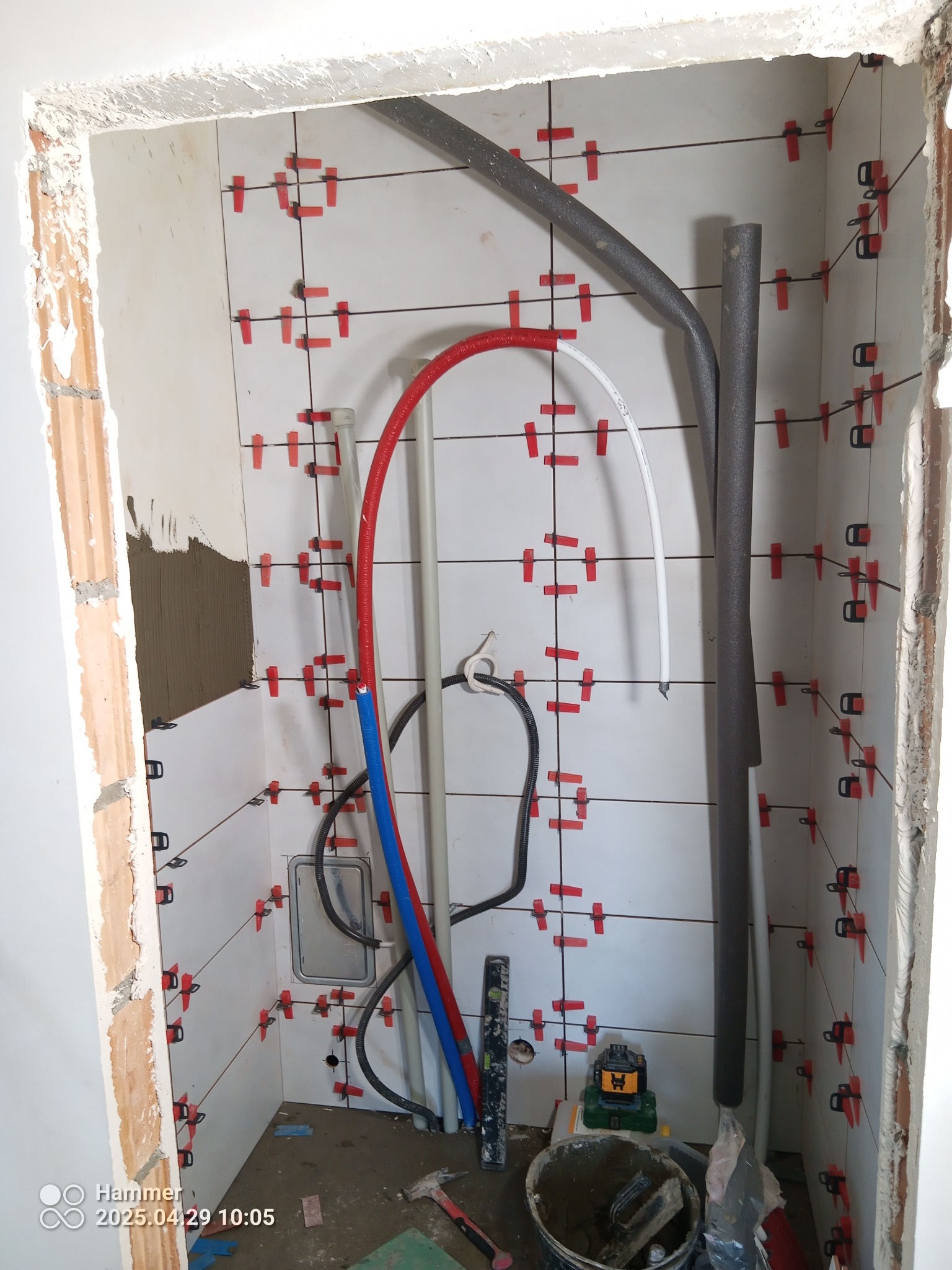 Białe płytki z czerwonymi krzyżykami dystansowymi na ścianie w trakcie prac. Widoczne rury, poziomica i wiadro z zaprawą. Fragment ściany z nałożoną zaprawą.
