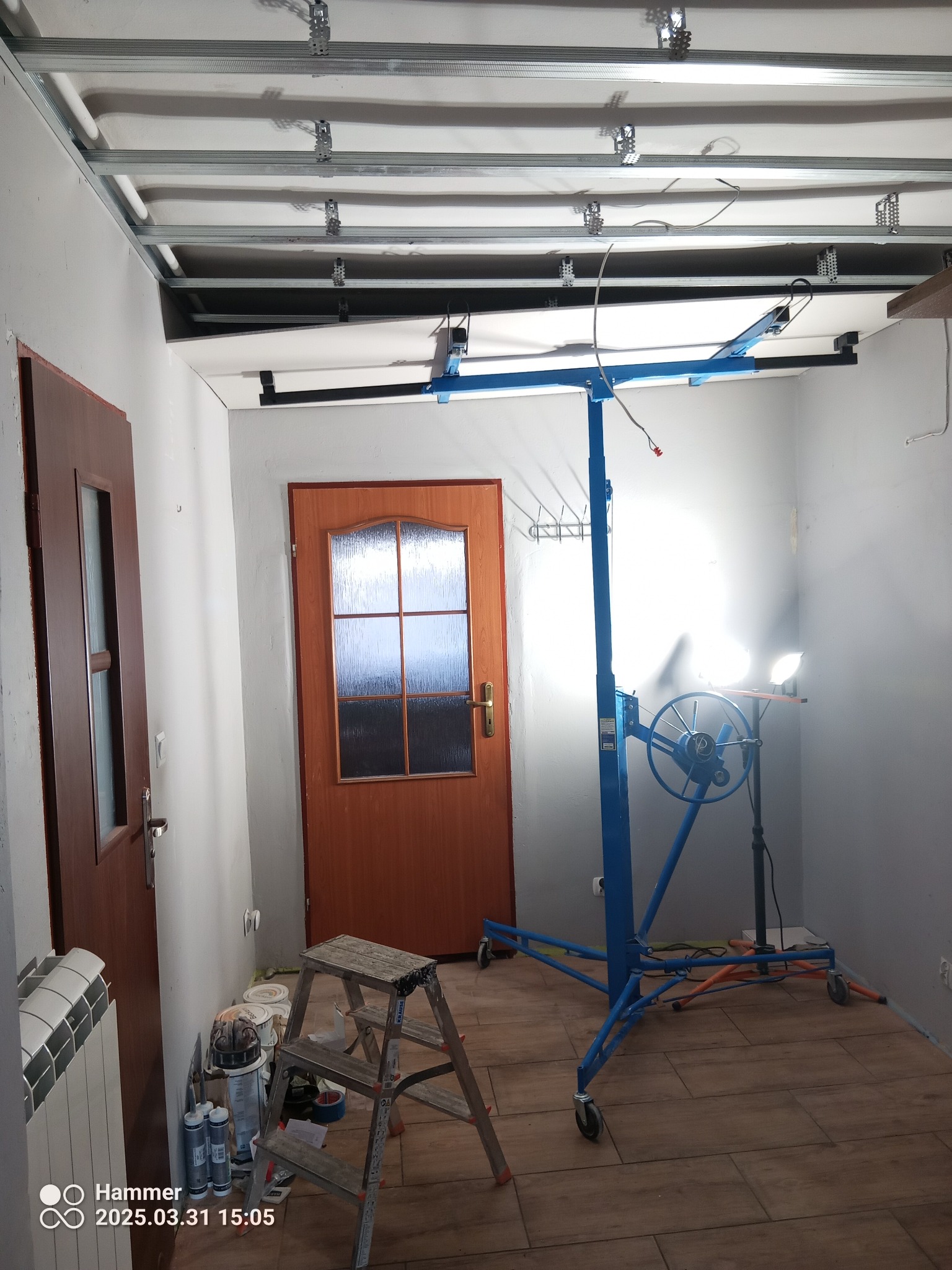 Montaż sufitu podwieszanego: metalowa konstrukcja, podnośnik do płyt, drabina, drzwi. Widoczne narzędzia i materiały. Oświetlenie robocze.