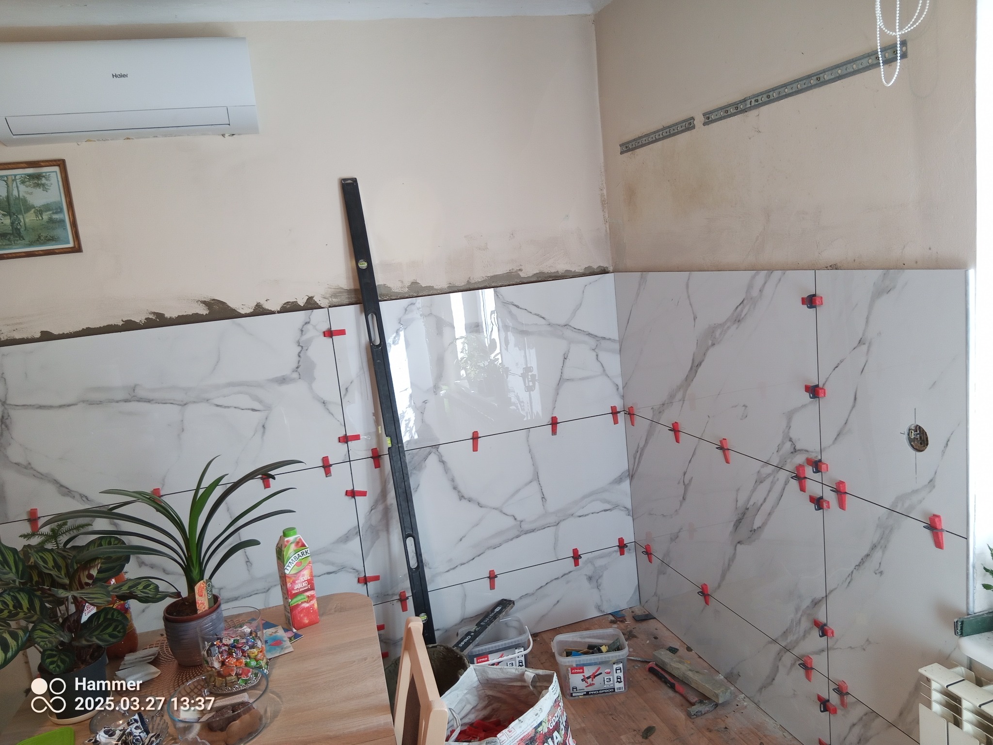 Ściana w trakcie układania płytek imitujących marmur, z użyciem poziomicy i czerwonych klipsów dystansowych. Widoczne narzędzia i akcesoria glazurnicze.