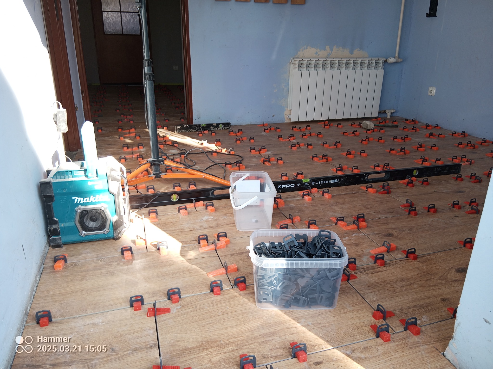 Brązowe płytki podłogowe w trakcie układania, z użyciem klipsów poziomujących i poziomicy. Widoczny grzejnik i sprzęt budowlany.