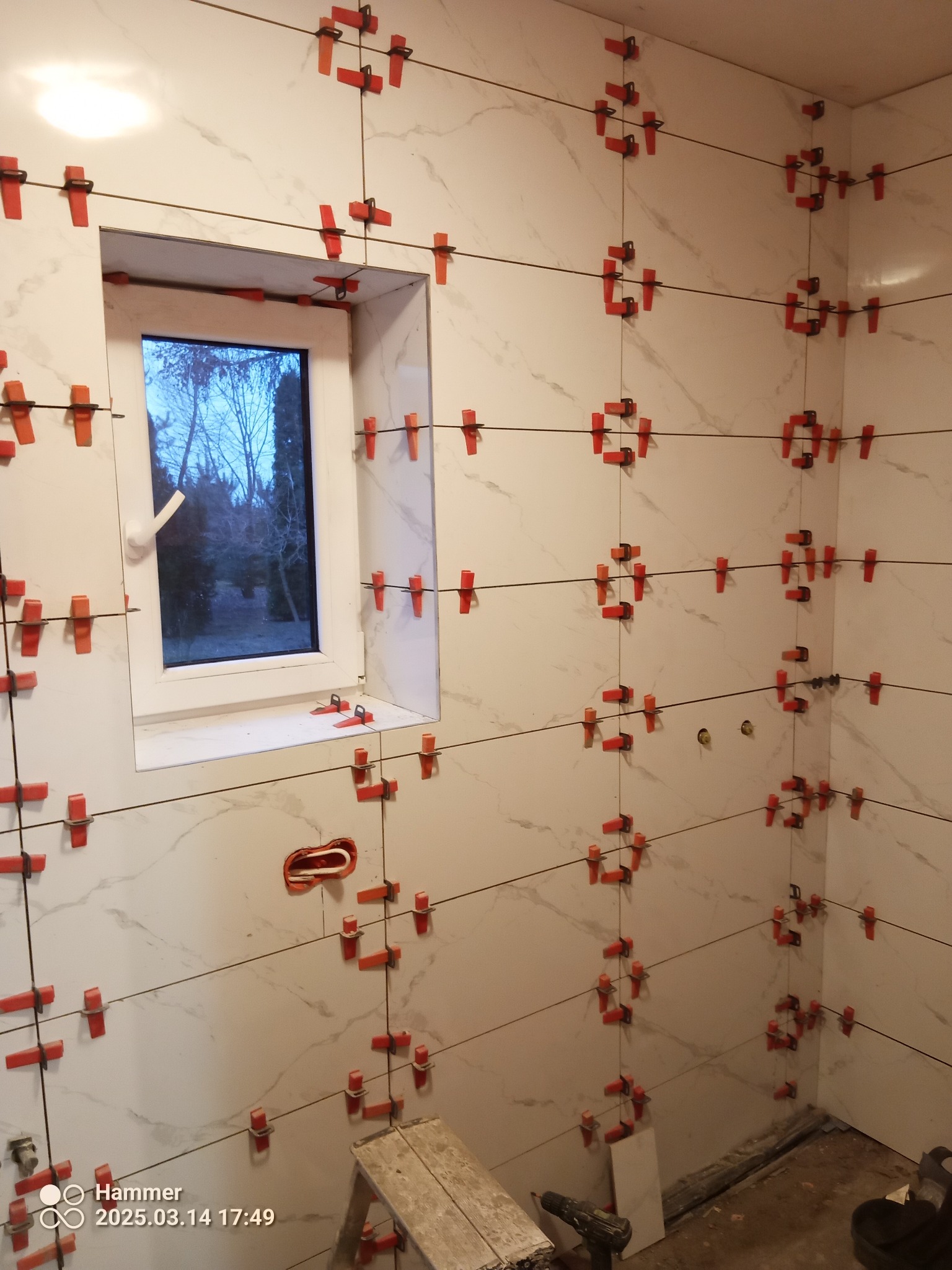 Ściana w trakcie układania płytek z użyciem systemu poziomowania. Widoczne czerwone klipsy i kliny, okno z widokiem na zewnątrz, drabina i wiertarka w dolnej części kadru.