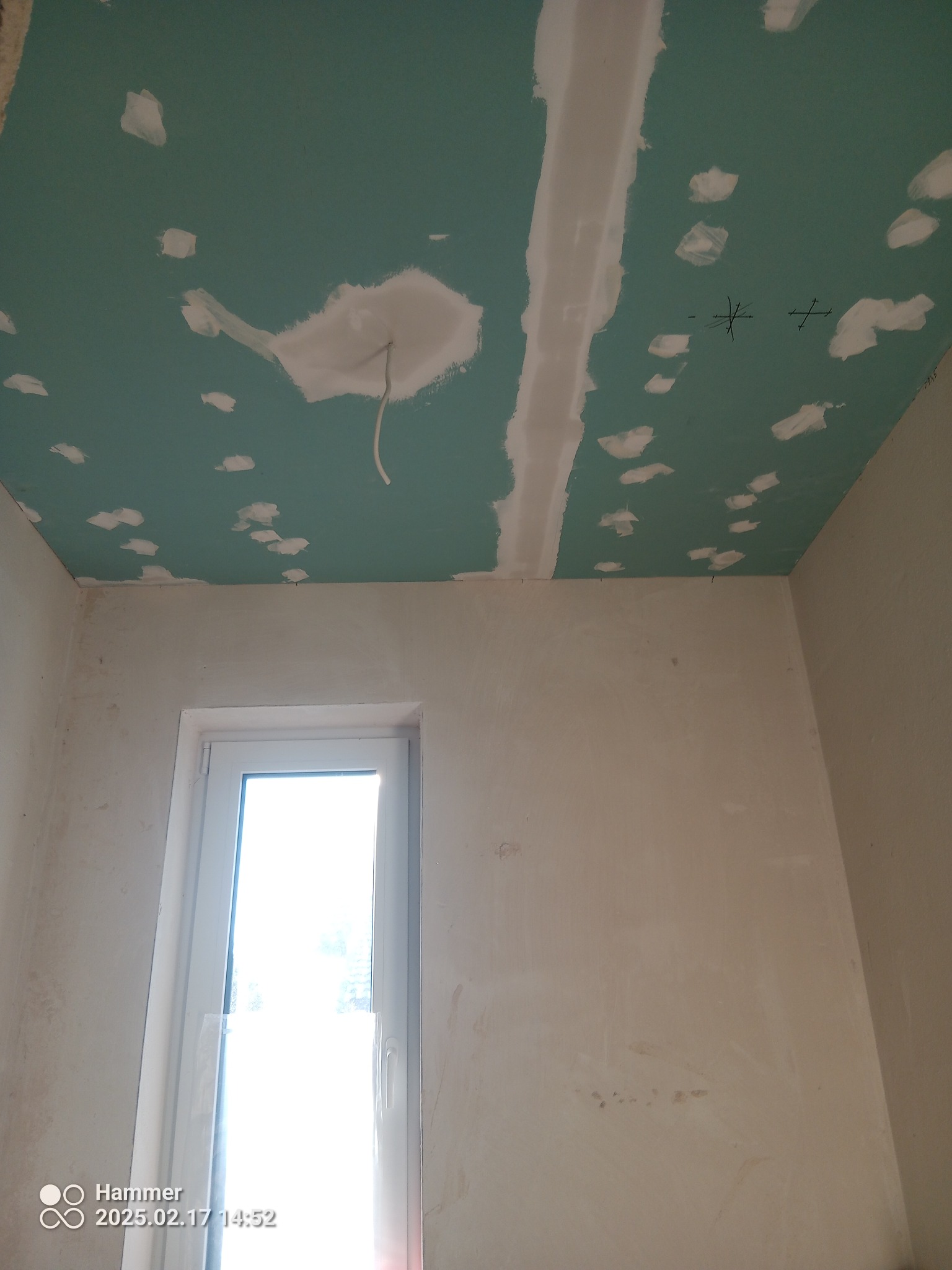 Nierówna, zielona płyta gipsowa na suficie po szpachlowaniu, z widocznymi łączeniami i miejscami po wkrętach. W tle ściana i okno z silnym światłem.