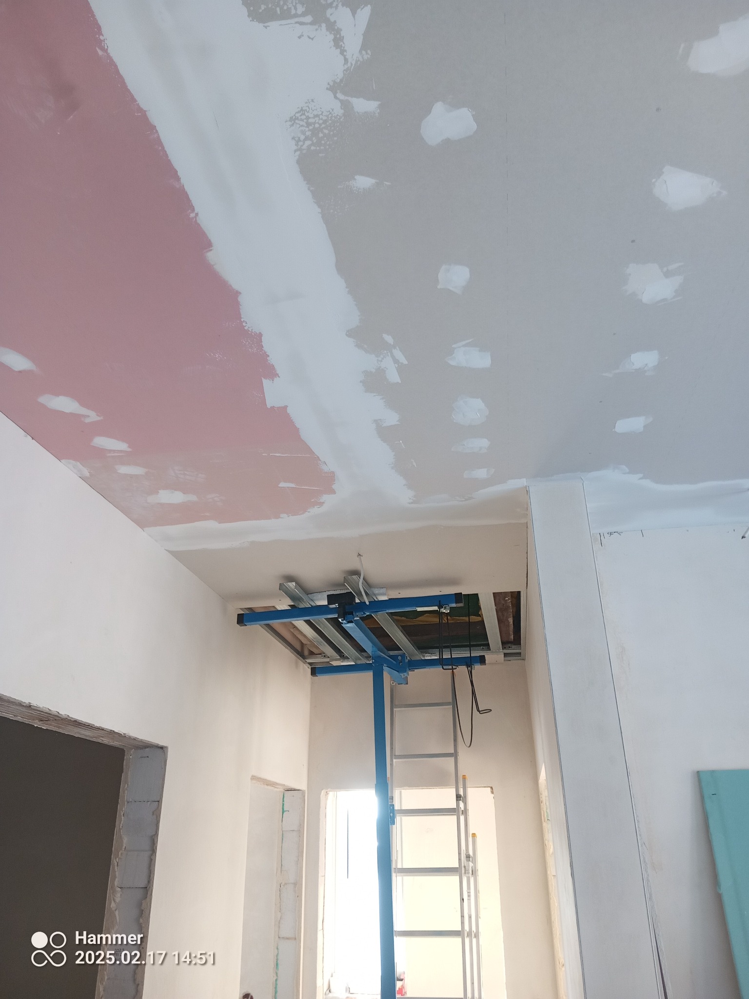 Sufit w trakcie prac: częściowo pomalowany na różowo, fragmenty z nałożoną gładzią, widoczny podnośnik do płyt gipsowych i drabina. W tle surowy otwór drzwiowy.