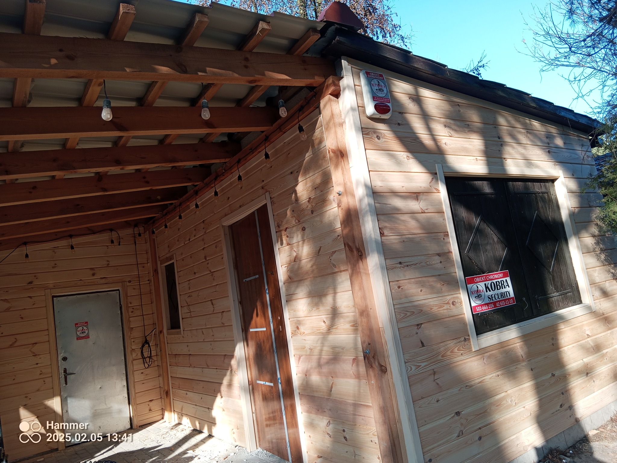 Drewniana konstrukcja z widocznym zadaszeniem, oświetleniem i drzwiami. Na ścianie tabliczka informacyjna o ochronie obiektu. Widoczne elementy wykończeniowe.