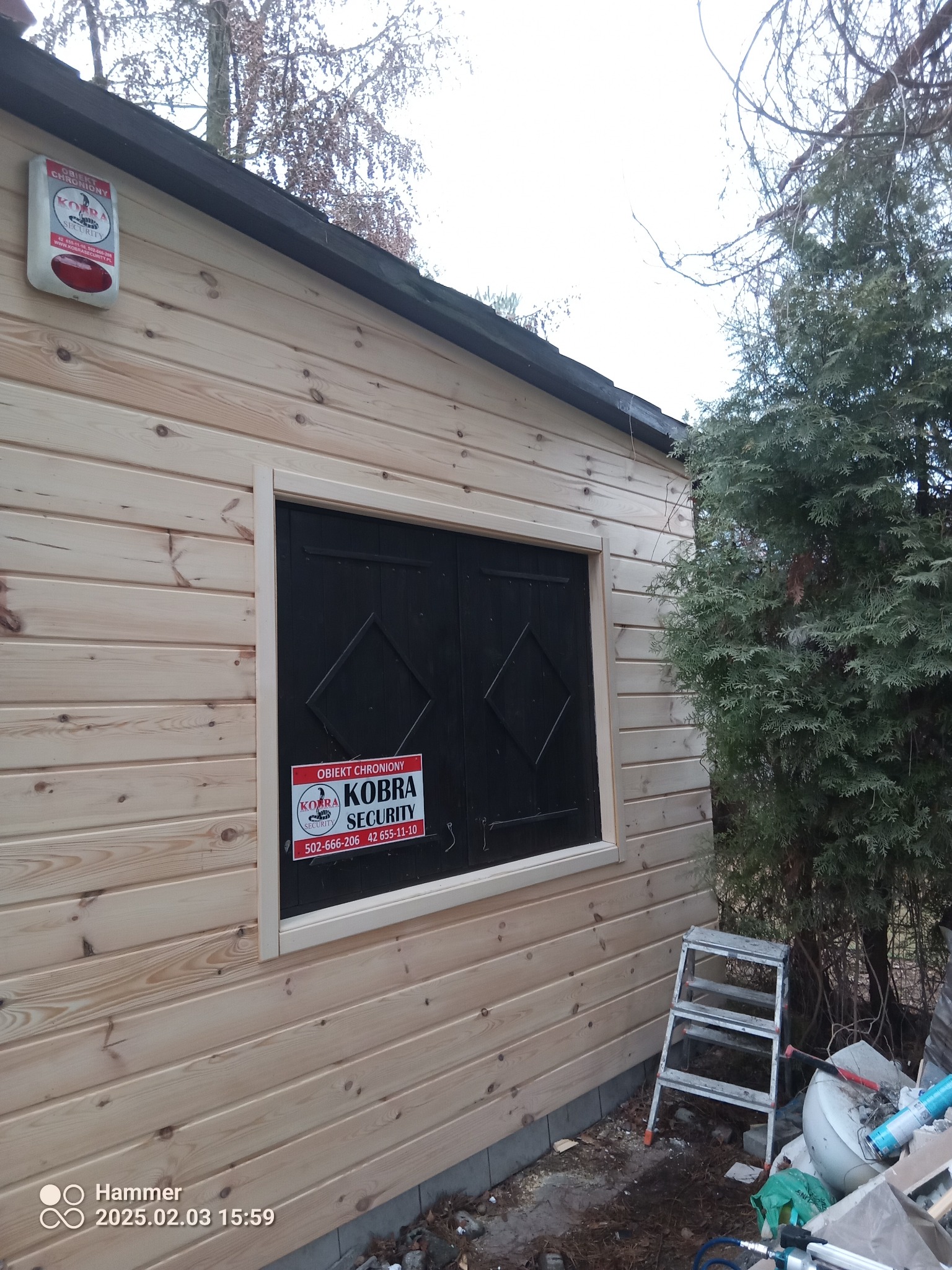 Drewniana konstrukcja z czarnymi okiennicami i tabliczką 'Obiekt Chroniony Kobra Security'. Widoczna drabina i elementy otoczenia. Ujęcie z dołu, lekko z prawej strony.