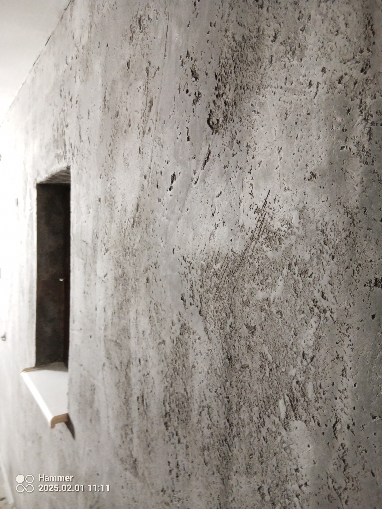 Szara ściana z efektem dekoracyjnym, widoczna wnęka okienna z białym parapetem. Tekstura ściany nierówna, z licznymi wgłębieniami i wypukłościami. Ujęcie z boku, blisko ściany.