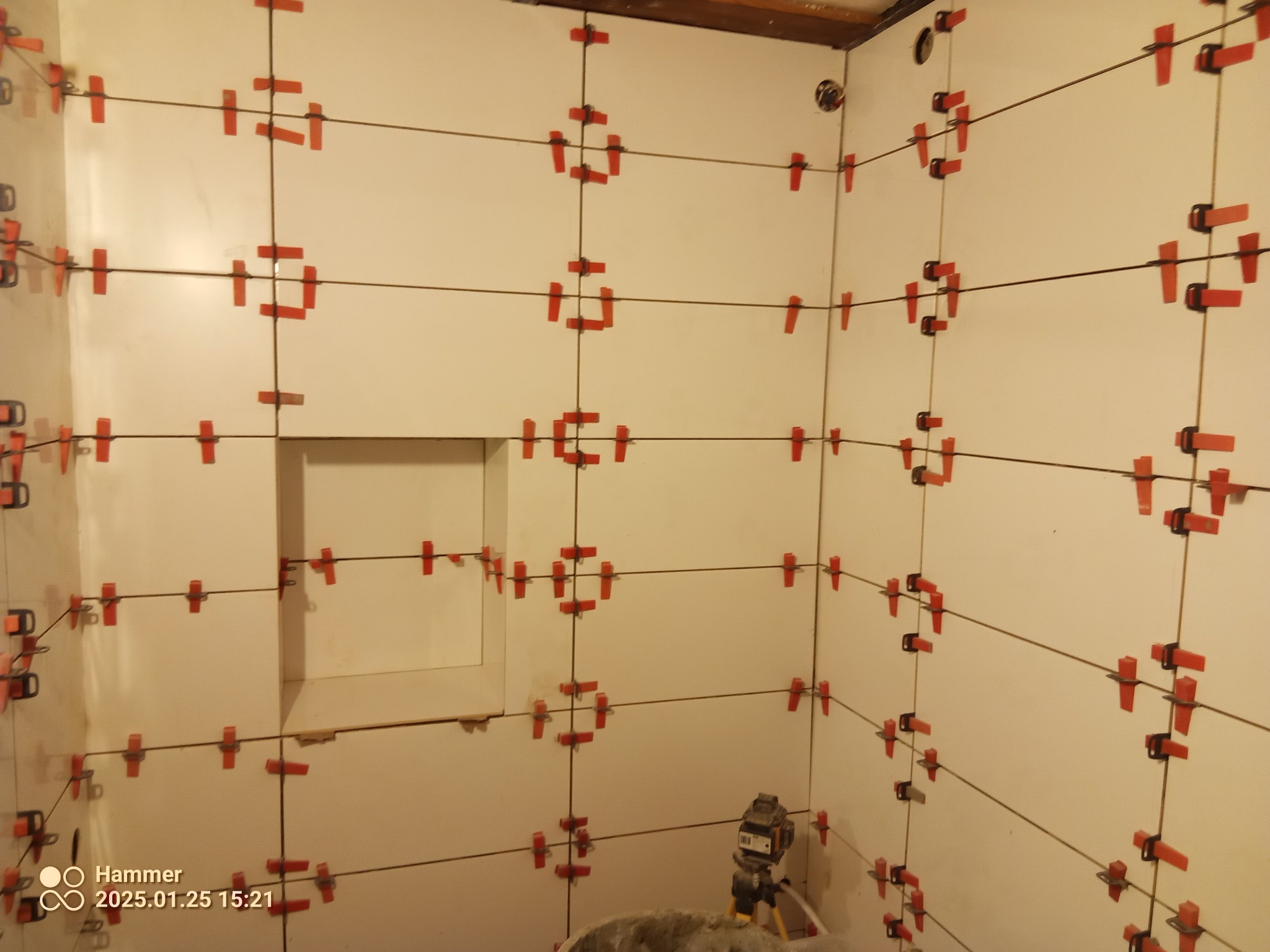 Białe płytki ścienne w trakcie montażu, z użyciem czerwonych klipsów poziomujących i lasera krzyżowego. Widoczny fragment ściany z wnęką.