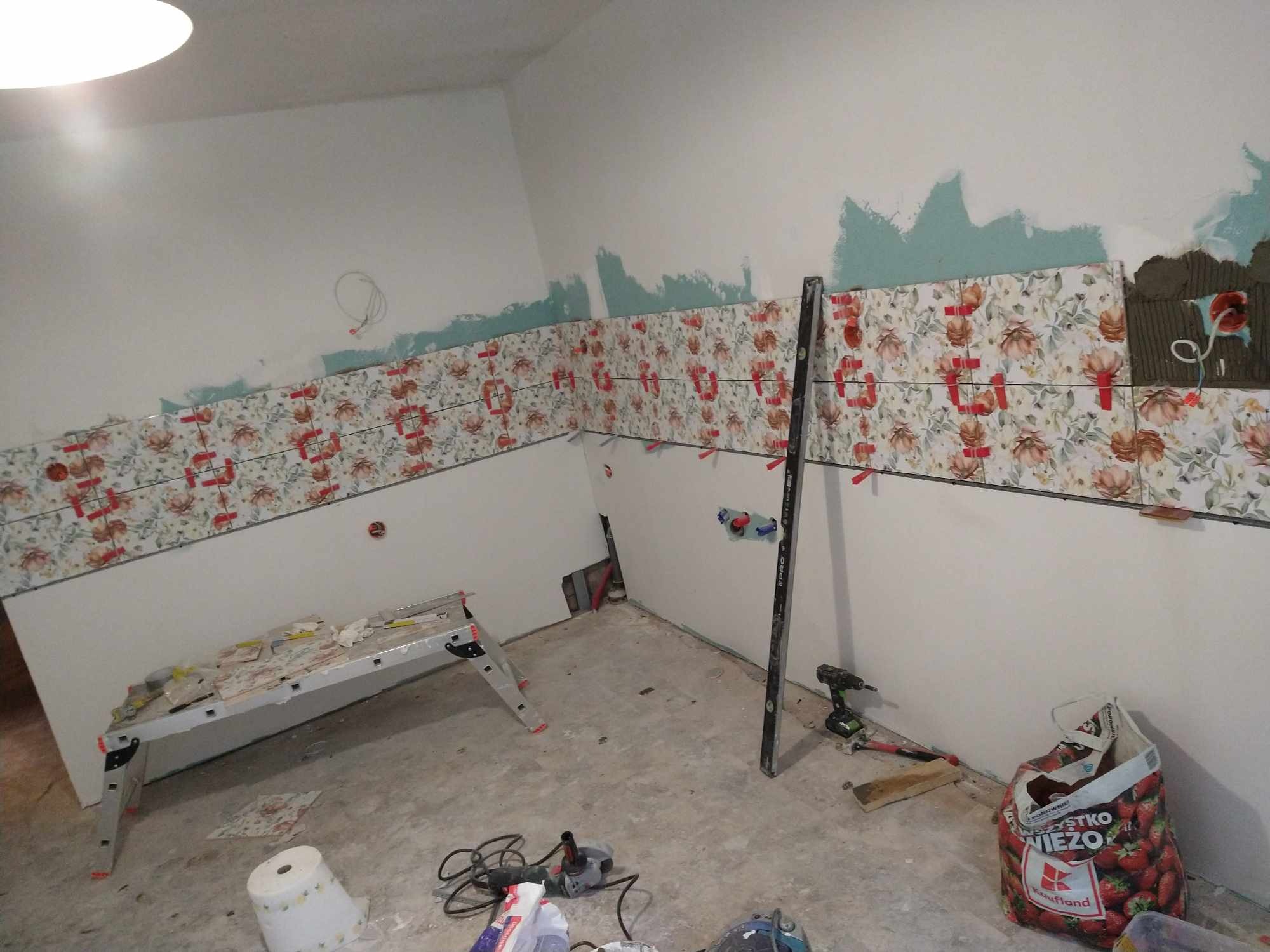 Układanie płytek z motywem kwiatowym na ścianie w kuchni. Widoczny poziomica, drabina i narzędzia glazurnicze. Prace w toku, fragment ściany z nałożonym klejem.