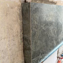 Mariusz Pala - Montaż płyty z efektem marmuru na ścianie z widocznymi śladami przygotowania podłoża. Widoczny profil montażowy i fragment instalacji wodnej.