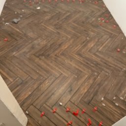 Mariusz Pala - Ukończona podłoga z płytek imitujących drewno, ułożonych w jodełkę, z czerwonymi krzyżykami dystansowymi. Widok z góry, fragment ściany i podłogi w tle.