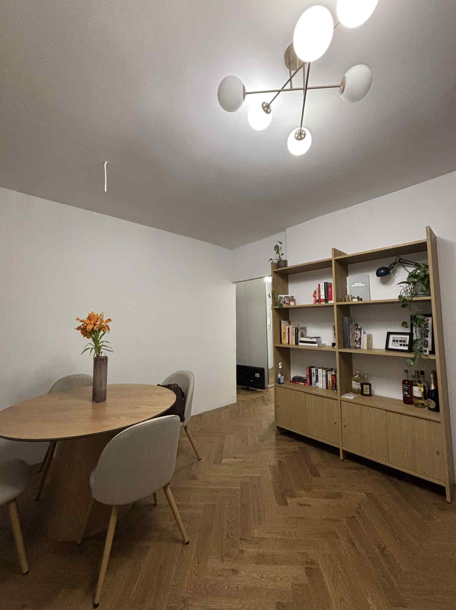 Jasne, parkietowe wnętrze z drewnianym regałem, okrągłym stołem i nowoczesną lampą sufitową. Minimalistyczny design z kwiatami w wazonie.