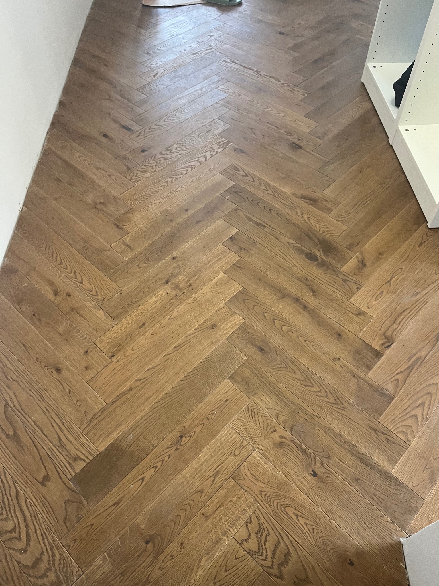 Ułożony parkiet w jodełkę francuską z widoczną strukturą drewna i naturalnym odcieniem brązu, częściowo widoczny biały regał w tle.