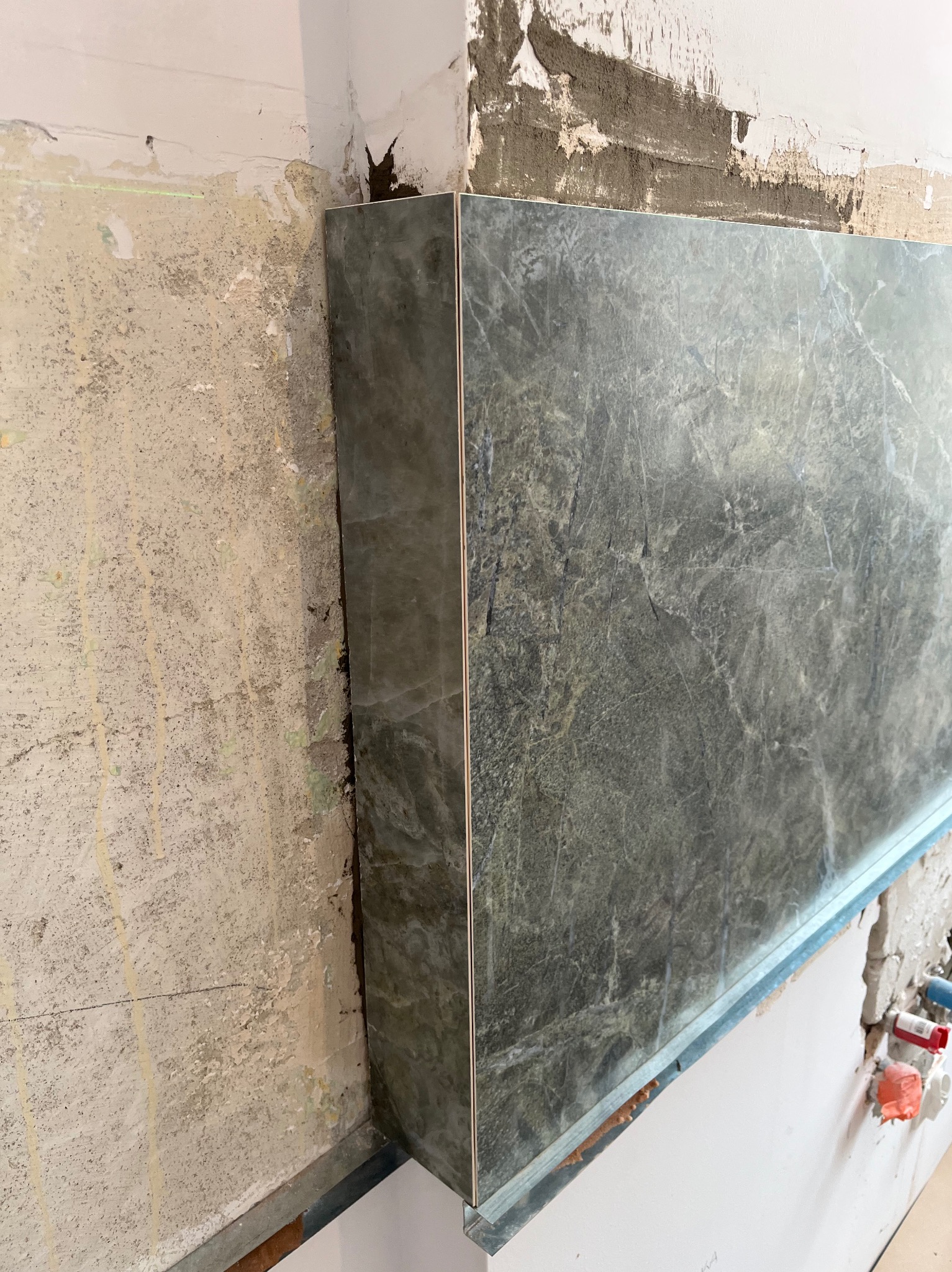 Montaż płyty z efektem marmuru na ścianie z widocznymi śladami przygotowania podłoża. Widoczny profil montażowy i fragment instalacji wodnej.