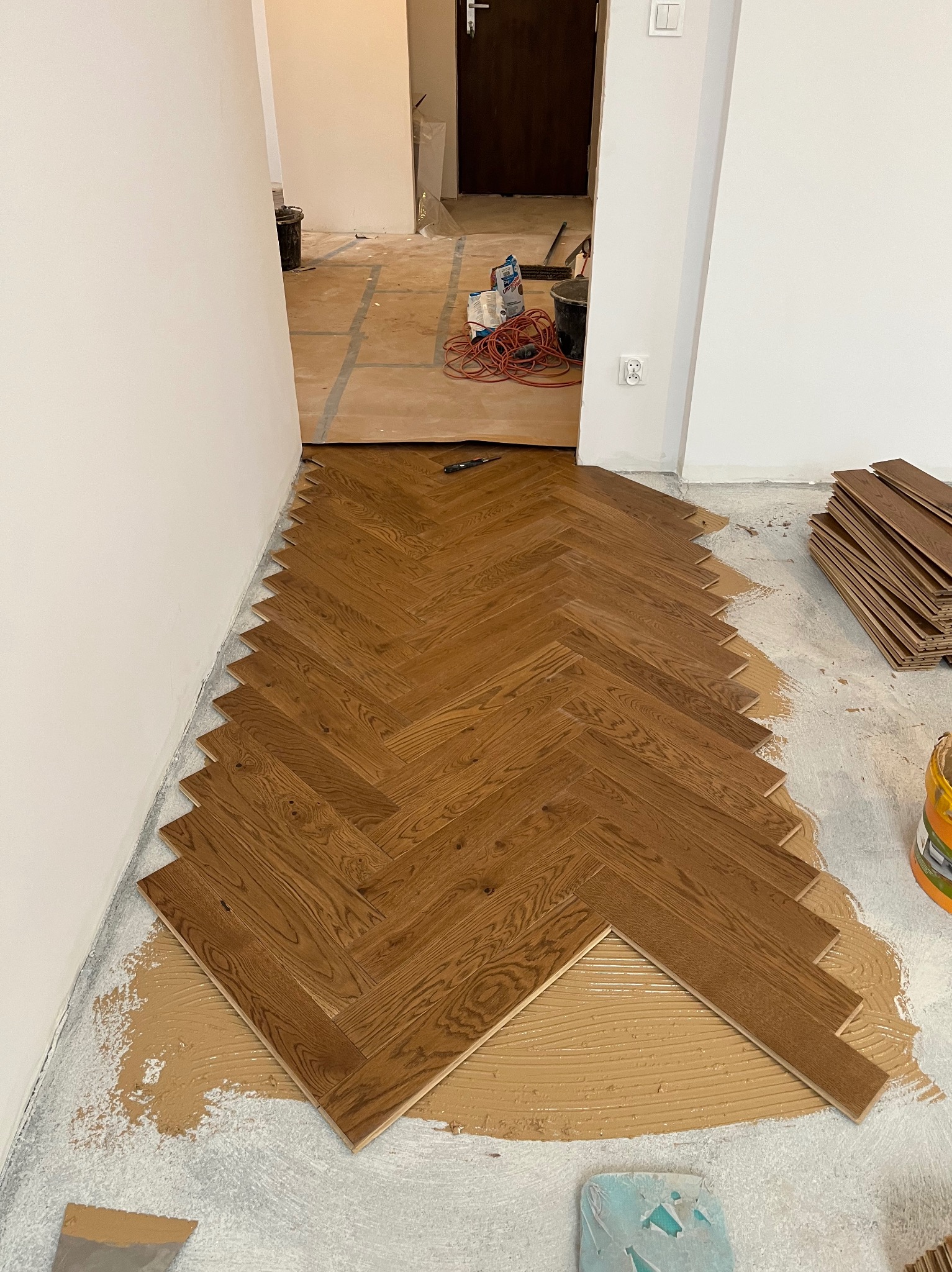 Układanie parkietu w jodełkę na kleju. Widoczne deski parkietowe, klej, narzędzia i przygotowane podłoże. Częściowo ułożona podłoga w trakcie prac wykończeniowych.