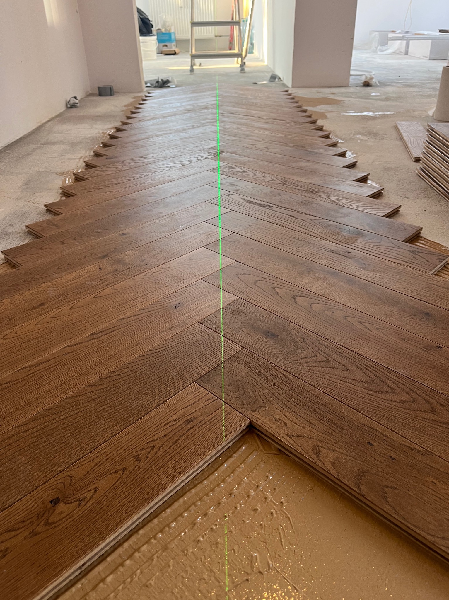 Układanie parkietu w jodełkę z użyciem lasera w Wiśniowej. Widoczny klej i fragmenty nieułożonych desek. Parkiet w ciepłym odcieniu brązu, surowe ściany w tle.