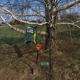 Lex Terrae Łukasz Szajewski - Sprzęt pomiarowy i odzież robocza firmy geodezyjnej zawieszone na brzozie na tle pola. Widoczna łopata i notatnik. Słoneczny dzień.